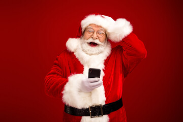 Cheerful Santa Claus in a red suit poses with a smartphone against a festive red background as he shares holiday joy and celebrates Christmas season