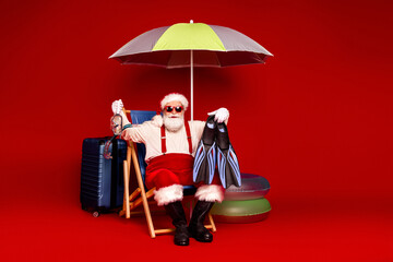 Santa Claus relaxes in a bright red studio with luggage an umbrella and fins preparing for...