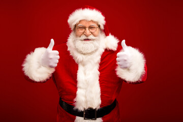 Santa Claus gives thumbs up in red suit with white fur trim smiling for holiday photo against red background