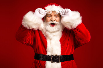 Santa Claus in red suit adjusts his glasses with a surprised smile against a solid red background ready for christmas season celebration