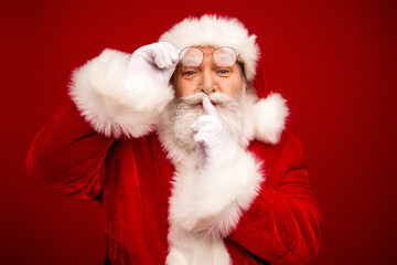 Santa with glasses and white beard in a red suit poses with finger to lips in a festive holiday portrait for Christmas season
