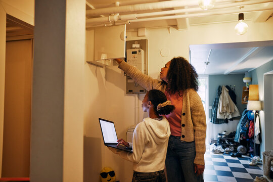 Mother checking electrical meter with daughter holding laptop while standing in basement