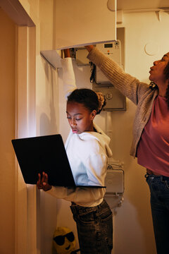 Girl checking wi-fi connection in laptop with mother near electrical meter at illuminated basement