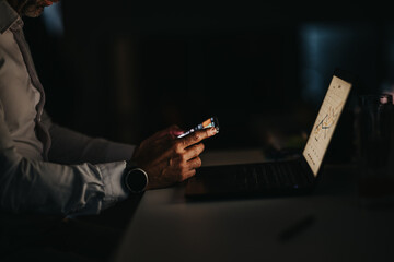 A focused businessman works at a dimly lit desk, scrolling on a smartphone while a laptop displays a chart. The scene conveys remote work, data analysis, and digital collaboration.
