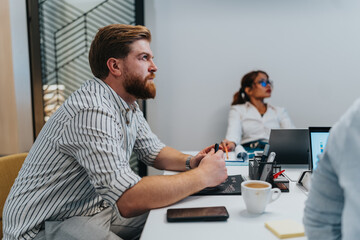 A bearded man sits at a conference desk, taking notes as colleagues work on laptops in a bright,...