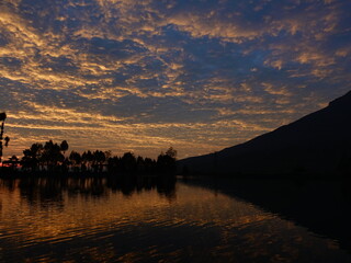 The dawn atmosphere with golden rays of sun in the blue sky, silhouettes of trees, and reflections on the lake water create a calming combination of views.