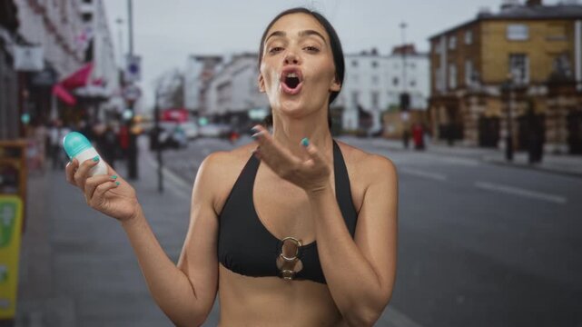 Woman blowing kiss to camera while holding a deodorant bottle on a busy city street; playful affection.