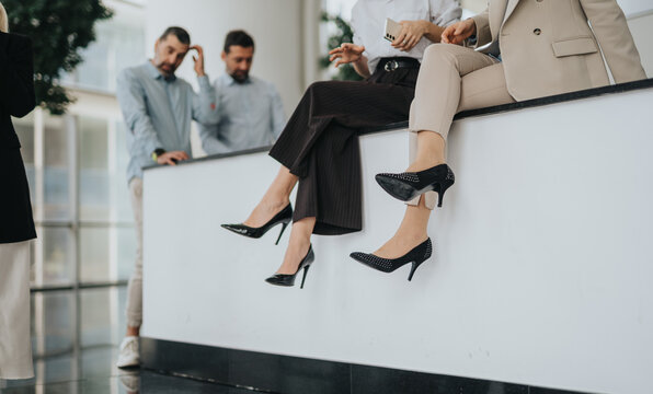 A group of professionals relaxes on a white ledge in a bright office lobby, dangling their legs in stylish heels. They appear focused yet at ease, pausing between meetings.