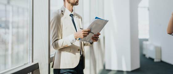A business professional in a beige suit holds a clipboard and paperwork in a bright office corridor, looking ahead.