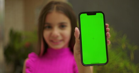 Young girl holding smartphone with green screen chroma key close to the camera, smiling warmly, showcasing a blank screen for customizable content in an outdoor home setting