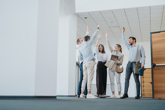 A diverse group of professionals in a modern office celebrate a success together, raising their hands in victory with smiles. They carry bags and devices, signaling teamwork and a positive workplace.