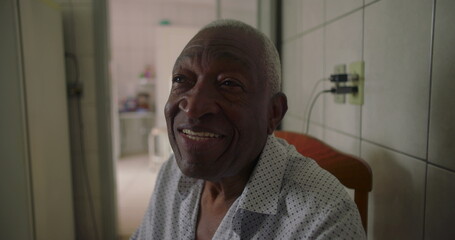 Elderly man of African descent sitting sideways in tiled kitchen, gently smiling while talking,...