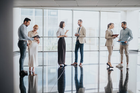 A diverse team of colleagues in smart attire stands in a bright glass-walled lobby, sharing ideas and notes during a casual business discussion. - Powered by Adobe