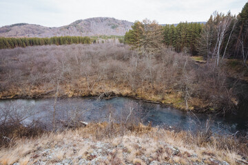 Calm river winds through fall terrain, Tranquil water flows amid autumnal foliage and hills, Gentle stream winds past arid shrubs and distant evergreen mountains under overcast sky