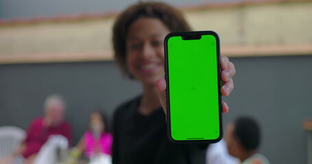 Teenager of African descent holding smartphone with green screen, chroma key display, smiling confidently, casual gathering with diverse adults in the background
