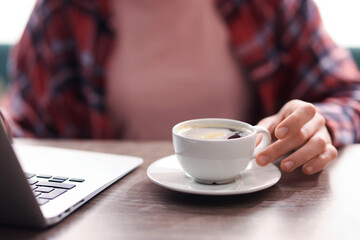 Coffee break. Woman with cup of aromatic drink and laptop at wooden desk indoors, closeup