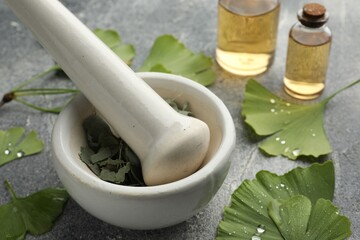 Wet ginkgo biloba leaves, mortar and bottles of essential oil on grey textured table, closeup