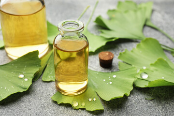 Wet ginkgo biloba leaves and bottles of essential oil on grey textured table, closeup