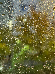 Close-up of raindrops on a window with blurred city lights and green trees in the background. Beautiful natural texture and rainy mood.