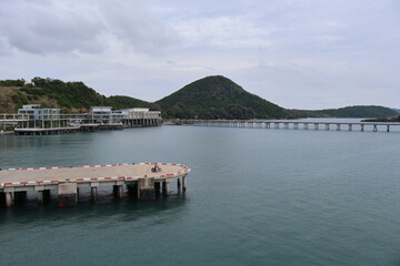 A tranquil harbor scene with calm waters reflecting the sky, a dock with piers stretching into the water, a hill with greenery in the distance, and an overcast sky.