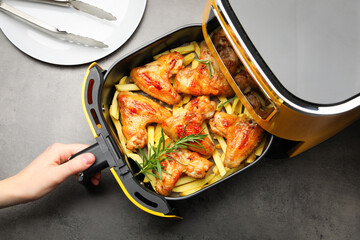 Woman opening air fryer with grilled chicken wings and french fries at grey table, top view
