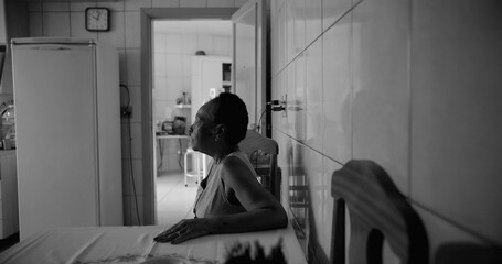 Elderly woman sitting at a dining table in a tiled kitchen, gazing thoughtfully towards the light,...