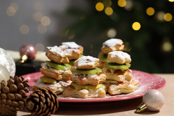 Sweet snacks made of puff pastry, slices of kiwi, cones and baubles on wooden table against blurred lights, closeup. Christmas food
