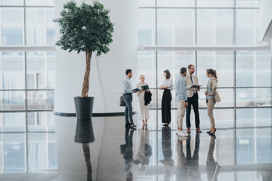 A diverse team of professionals stands in a bright, glass-walled lobby, exchanging ideas and reviewing files.