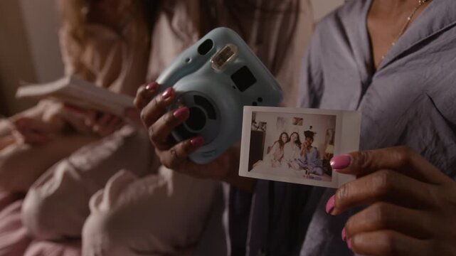 Medium midsection shot of hands of unrecognizable young black girl, wearing blue pajama, holding instant camera and photo with girlfriends during fun pajama party