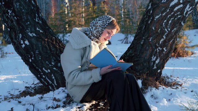 A beautiful young left-handed girl romantically admires the winter forest and writes something down in her notebook. Winter walk and creativity in nature. Healthy lifestyle.
