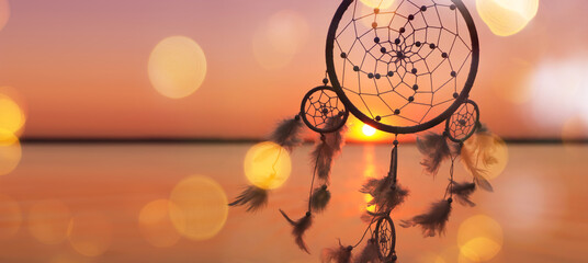 Dream catcher with feathers against sea at sunset, bokeh lights. Banner design
