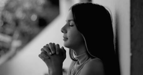 Young girl leaning against a wall, hands clasped in prayer, radiating hope and inner peace,...