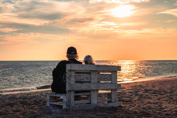 Two elderly people sit on a rustic wooden bench on a sandy beach, gazing at the calm ocean under a...