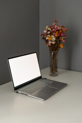 A sleek laptop sits open on a simple desk, paired with a vase of colorful dried flowers. This cozy workspace is bright and inviting