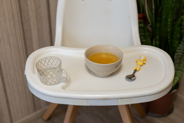 A bowl of soup sits on a high chair tray alongside a small glass and a colorful spoon, creating an inviting scene for a child's mealtime