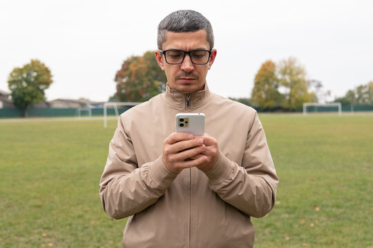 A man dressed in a light jacket stands on a soccer field, focusing on his smartphone while the sky is cloudy and trees are visible in the background