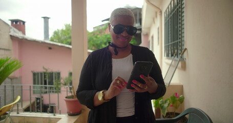 African American woman with short blonde hair and sunglasses smiling while using a smartphone on a balcony, surrounded by plants in a casual outdoor setting