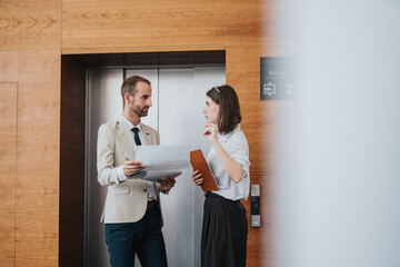 Two professionals in a contemporary office building stand near an elevator, reviewing papers and a...