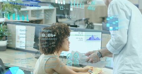 Seated woman and standing coworker reviewing financial charts on monitors in office, with documents