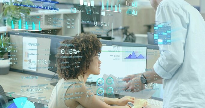 Seated woman and standing coworker reviewing financial charts on monitors in office, with documents