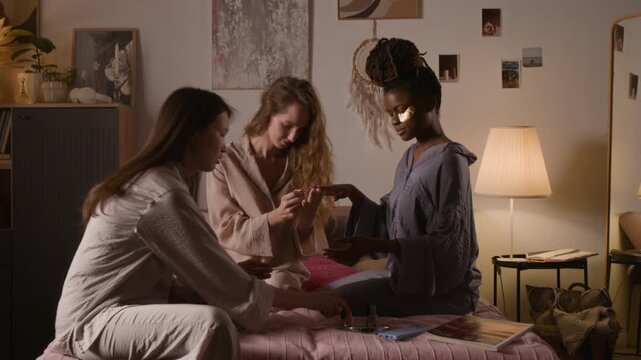 Full shot of three girlfriends, dressed in pajamas, sitting on double bed while having slumber party. African American girl getting manicure from Caucasian friend, Chinese woman showing off varnish