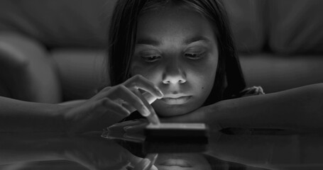 Close-up of young girl leaning on table, smartphone glow highlighting detached focus, capturing...