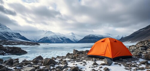 Vibrant orange tent contrasts rugged rocks and icy lake, snow peaks loom under cloudy winter sky,  nature,  solitude