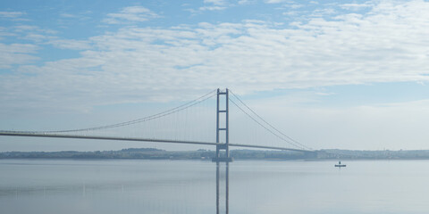 Wide, clear shot of a large suspension bridge, Humber Bridge with its main towers, spanning a wide river or estuary under a lightly cloudy blue sky.