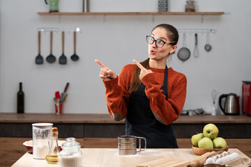 Woman in apron and eyeglasses pointing fingers left, showing client info while cooking food in the...