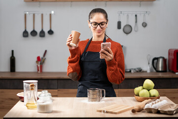 Woman housewife in apron and eyeglasses resting, using smartphone for social media and drinking takeaway delivery paper cup coffee after cooking food in the kitchen at home