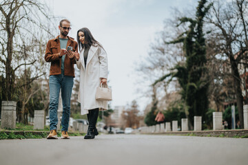 Fototapeta premium A man and woman walking outdoors in a city street, interacting with a phone. They appear relaxed and engaged, enjoying casual stroll in an urban setting surrounded by trees and buildings.