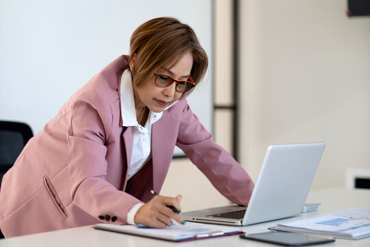 Asian adult businesswoman analyzing reports on laptop computer at office workspace professional diligent focused work desk