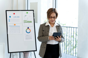 Asian adult businesswoman analyzing financial data reports on digital tablet device standing near whiteboard presenting market strategy charts office workspace