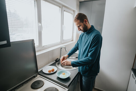 A man in a contemporary kitchen setting preparing eggs by a window with natural lighting.
